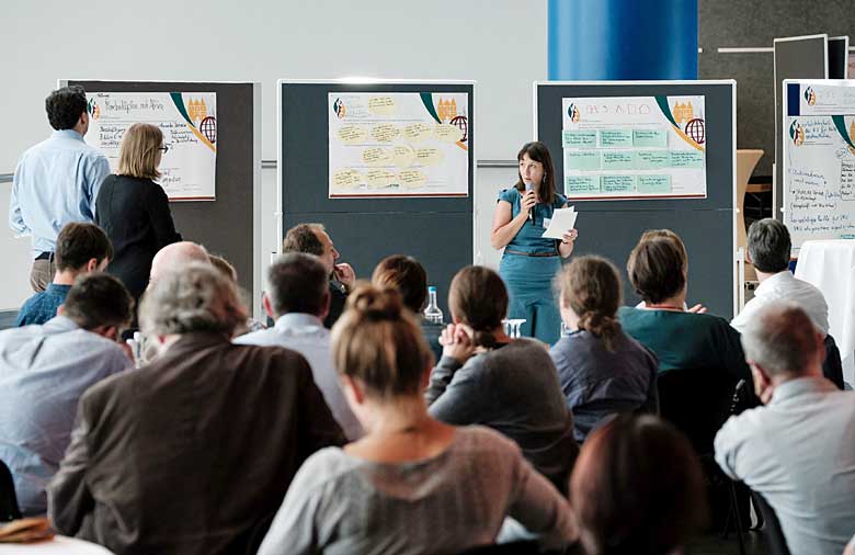 Eine Frau steht vor einem Publikum, spricht in ein Mikrofon und blickt dabei auf zwei Personen, die die neben ihr befindlichen Pinnwände beschriften.